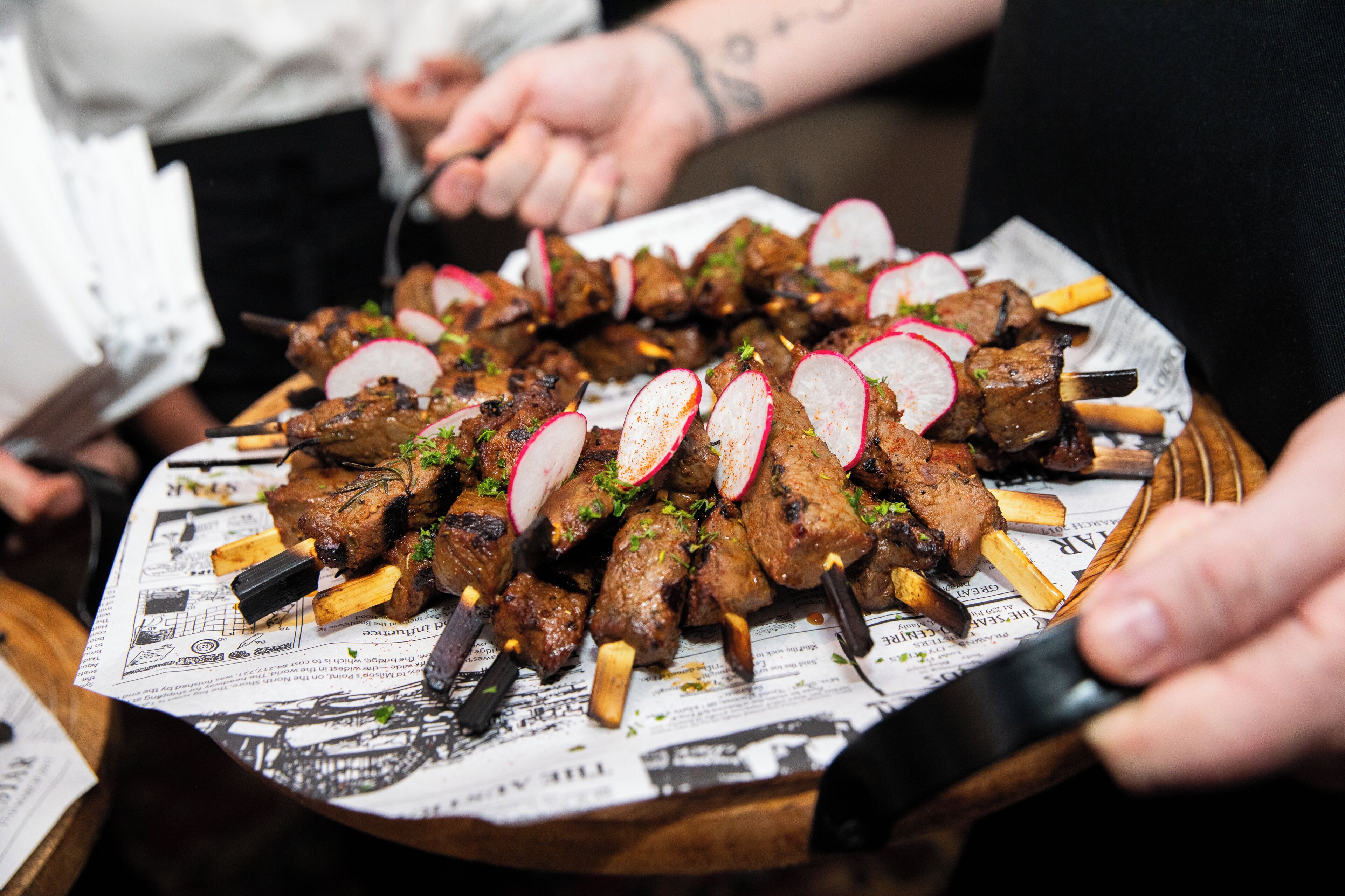 A platter of skewered meat with radish slices and herbs, served on a newspaper-lined tray.