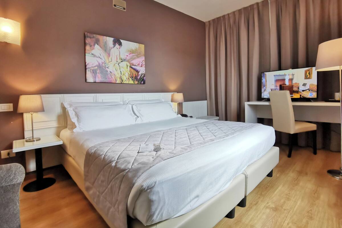 Una habitación de hotel con una cama grande, dos mesitas de noche con lámparas, un escritorio con silla, un televisor y un cuadro en la pared.