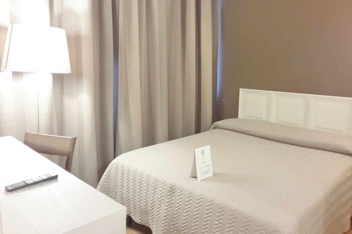 Une chambre d’hôtel avec un lit, un bureau, une chaise, une lampe et un panneau sur le lit.