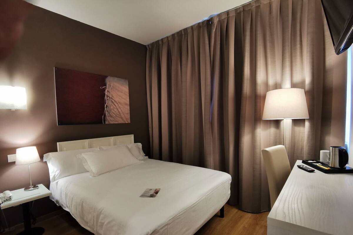 Habitación de hotel con una cama bien hecha, un escritorio con lámpara, una silla y un ventanal con cortinas.