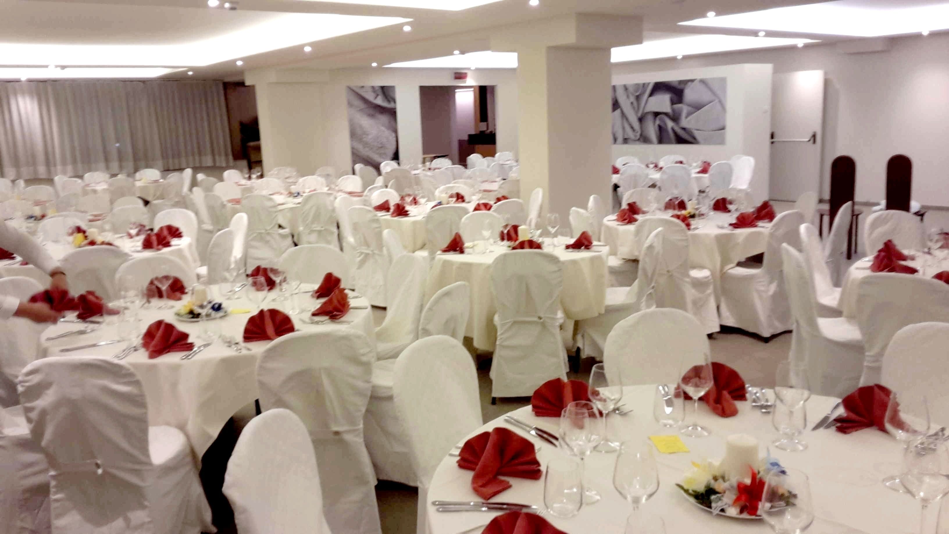 A banquet hall with round tables covered in white tablecloths and red napkins, arranged in rows.