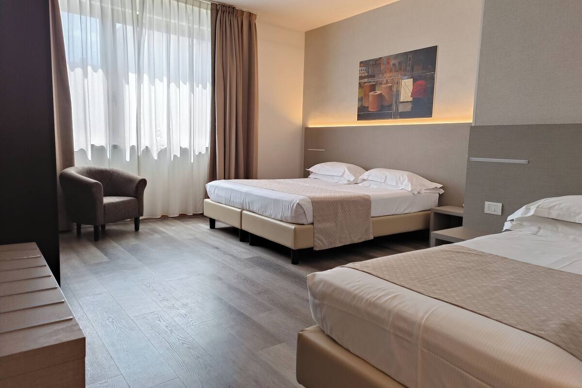 Habitación de hotel con dos camas, una silla, un cuadro en la pared y un ventanal grande con cortinas.