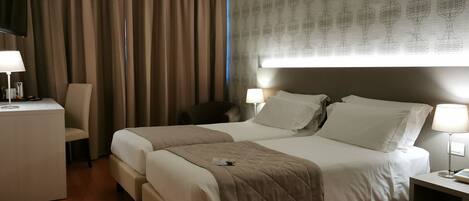 Habitación de hotel con cama doble, dos mesitas de noche con lámparas, un escritorio con lámpara, una silla y un ventanal con cortinas.