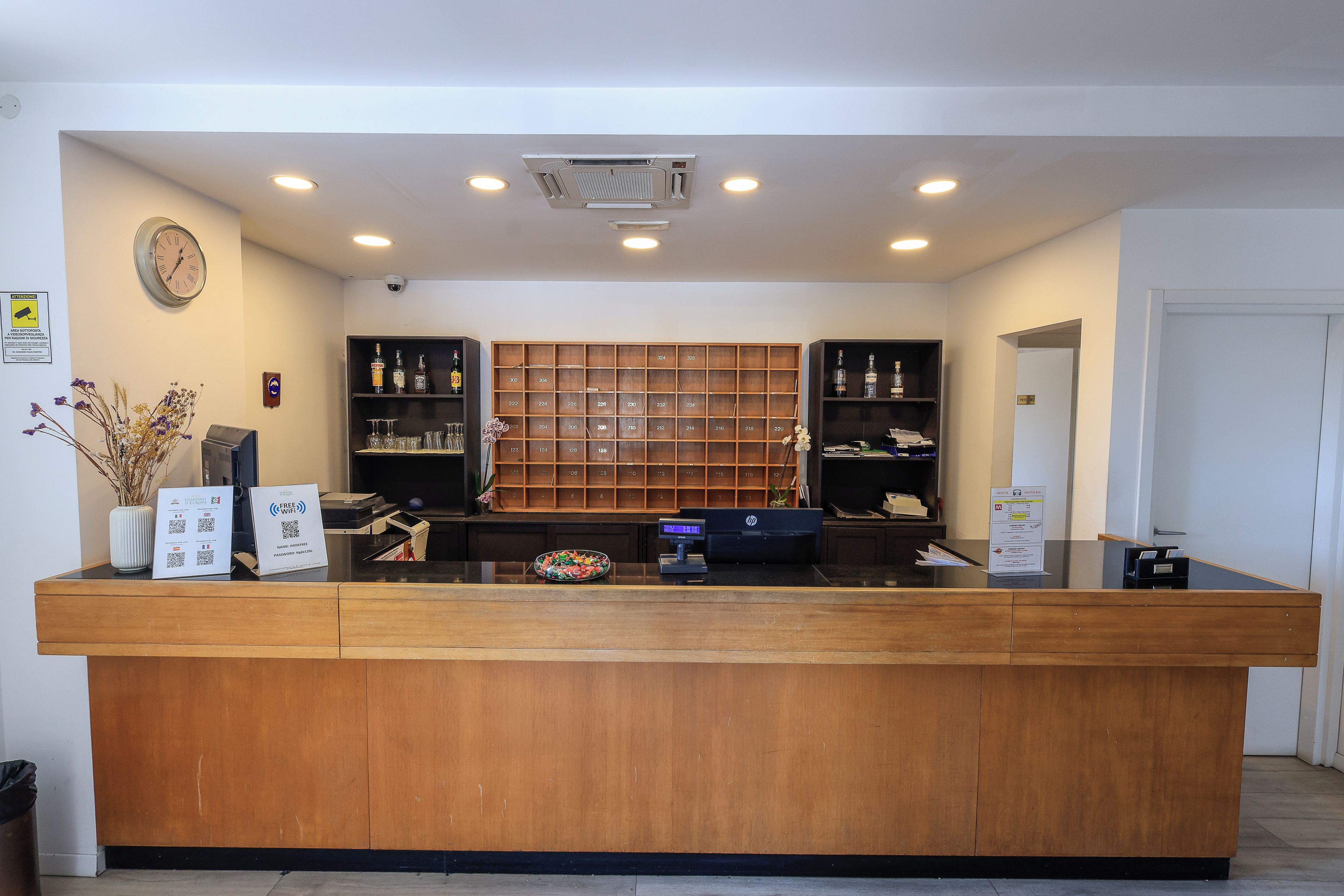 A hotel reception desk with a wooden counter, a clock on the wall, and a security camera.