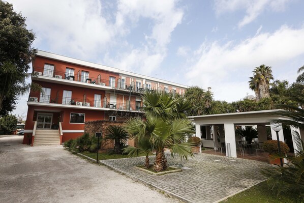 A multi-story building with a red facade, a covered outdoor seating area, and a paved courtyard with palm trees. - Hotel Giardino D'Europa (Rome)