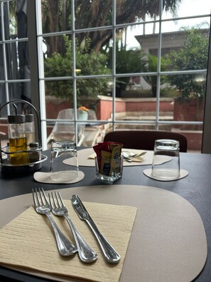 Une table dressée pour le repas, avec couverts, verres et condiments, donnant sur un jardin par une fenêtre.