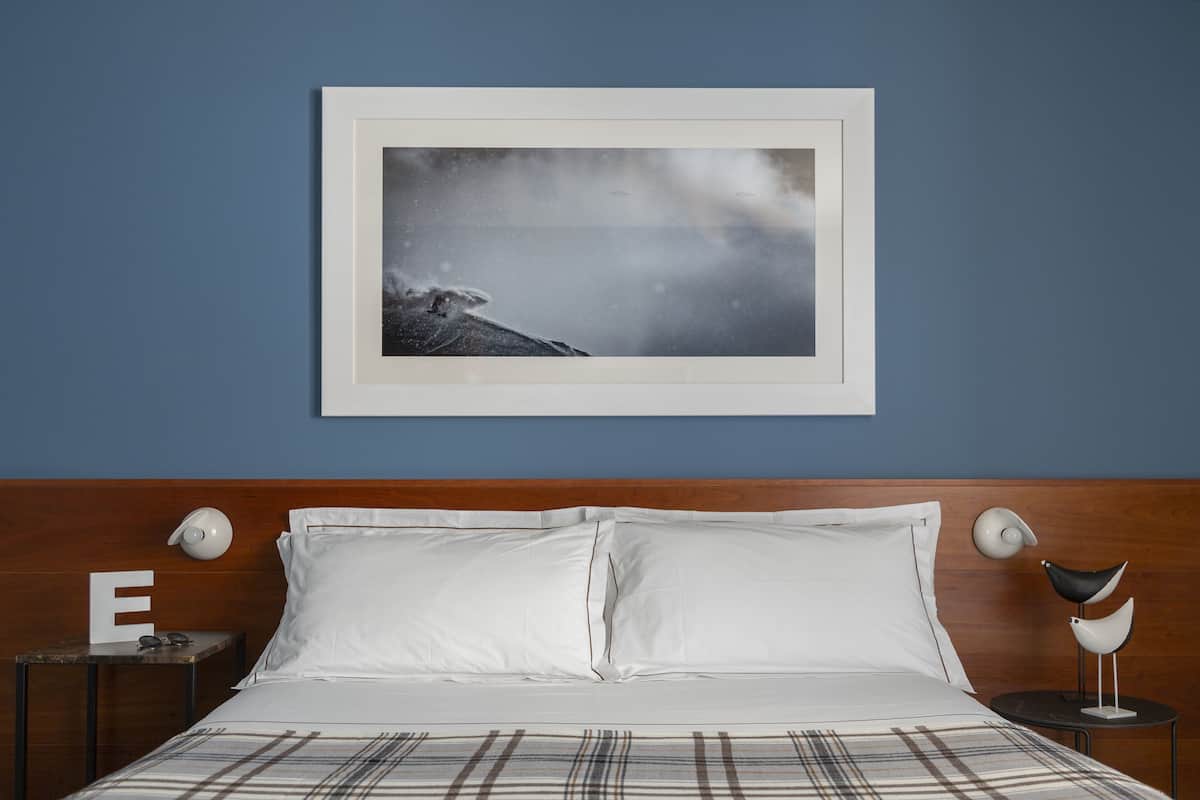 A neatly made bed with white linens, a plaid blanket, and a framed picture of a mountain landscape on the wall.