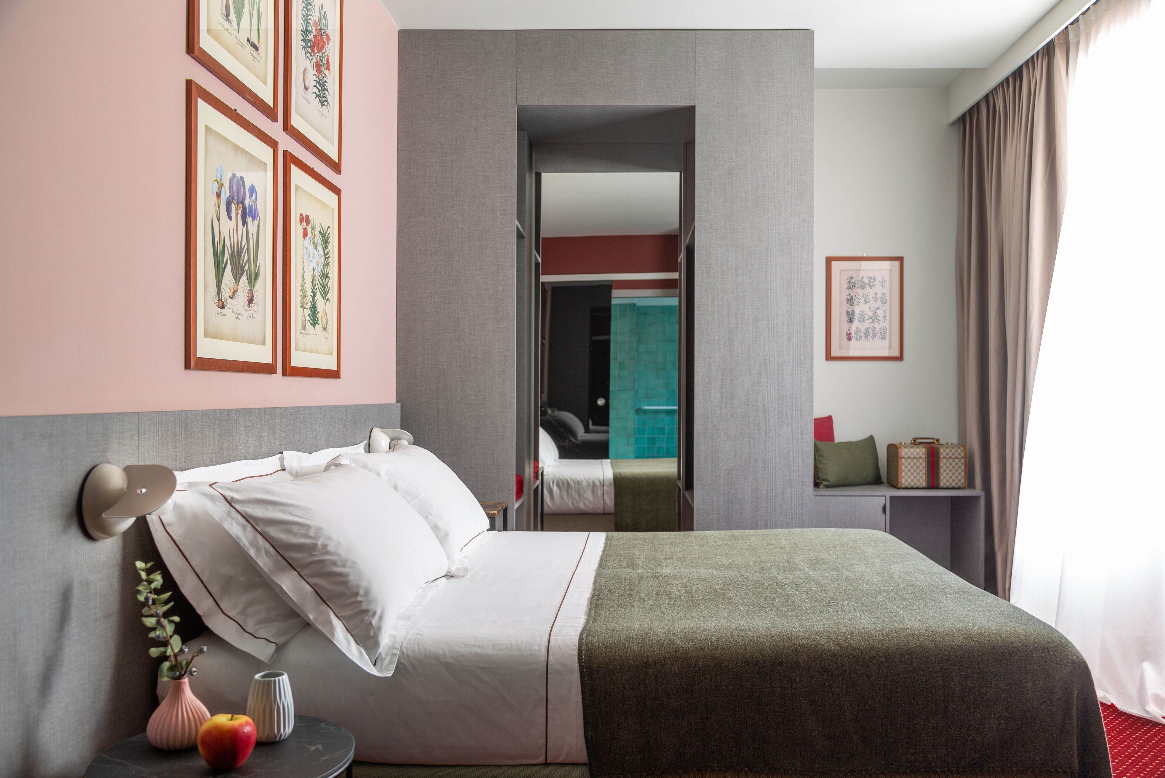 A hotel room with a bed, a nightstand with a vase and an apple, a mirror, a framed botanical print, and a view of the bathroom.