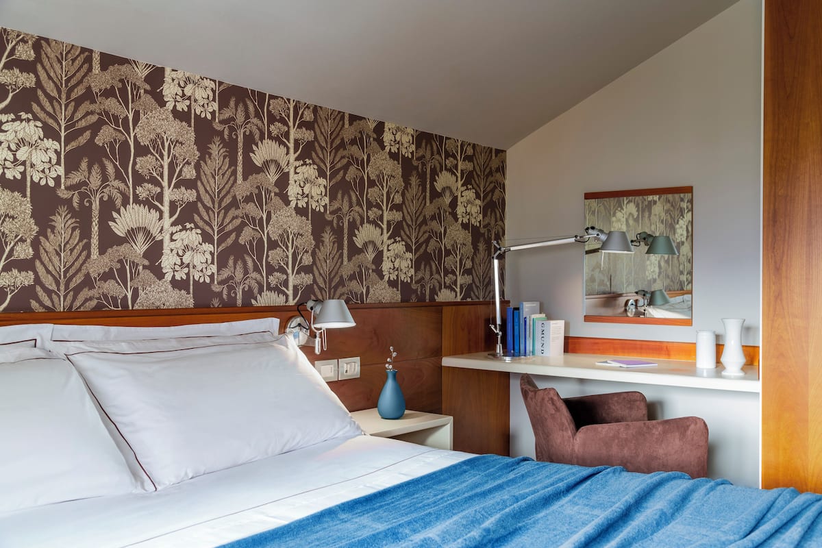 A hotel room with a bed, bedside table, lamp, chair, and a desk with a mirror and books.