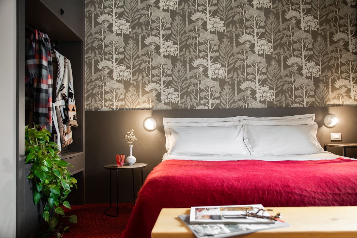 A hotel room with a bed, a nightstand, a small table with a vase, and a plant.