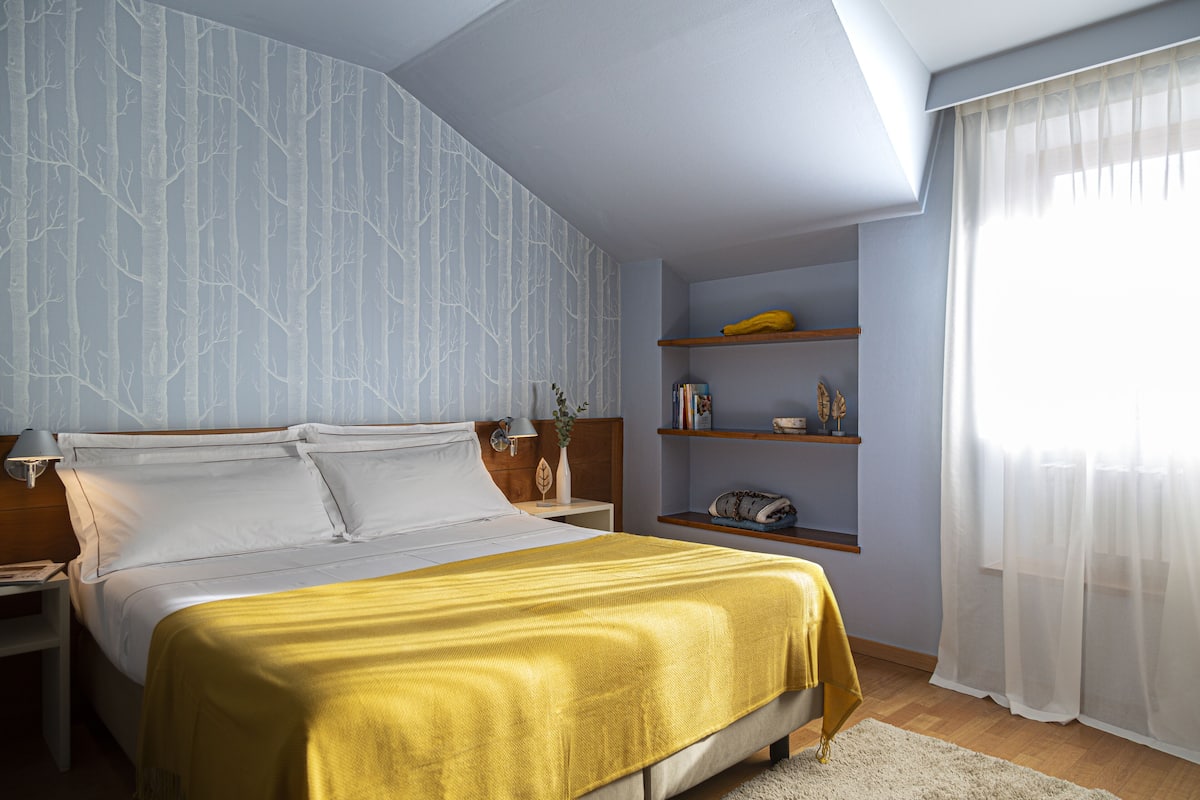 A bedroom with a bed, nightstands, a shelf with books and a banana, and a window with sheer curtains.