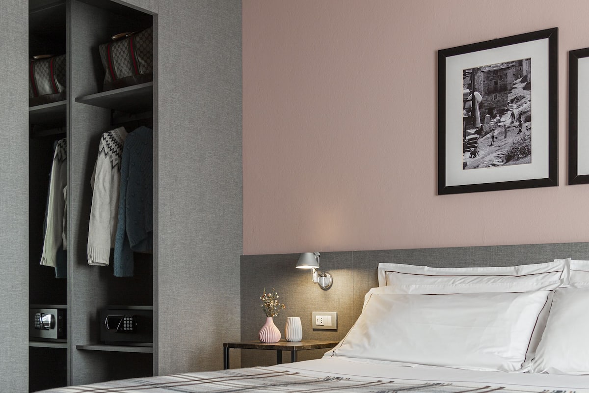A modern hotel room with a large bed, a bench, a wardrobe, and a bedside table with a lamp and a vase.