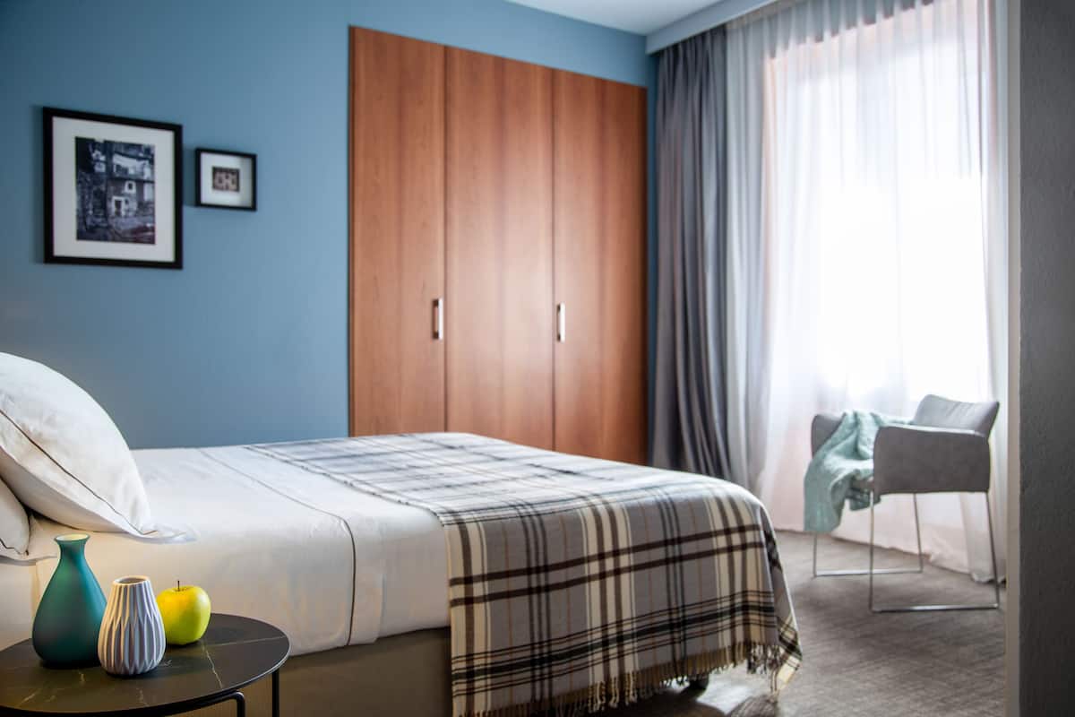 A bedroom with a bed, a wardrobe, a chair, and a table with a vase and an apple.