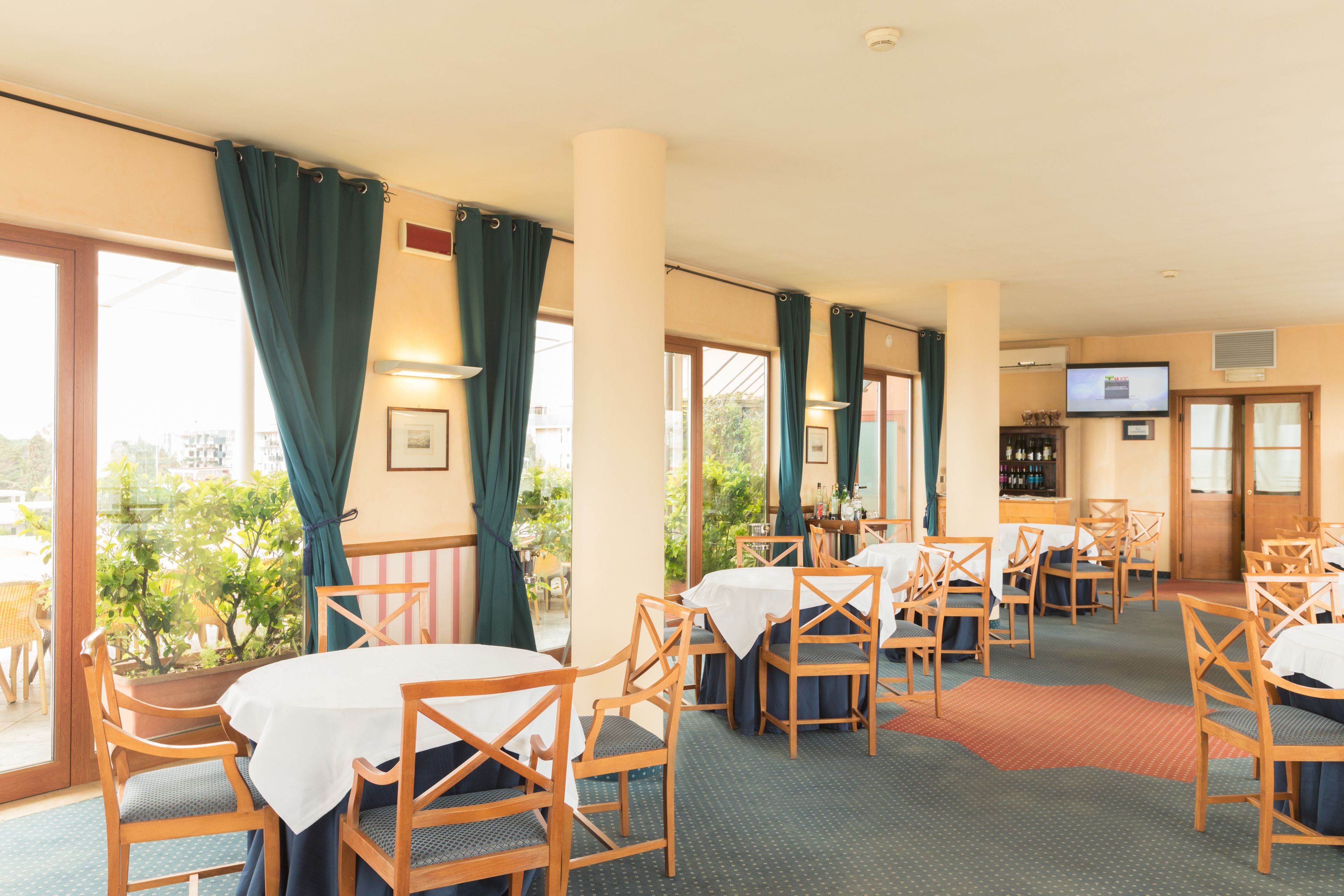 A hotel dining area with tables and chairs, large windows with curtains, and a television on the wall.