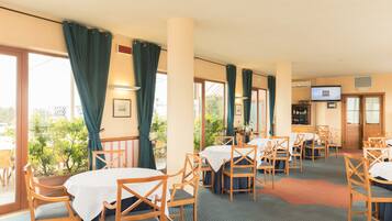 A hotel dining area with tables and chairs, large windows with curtains, and a television on the wall.
