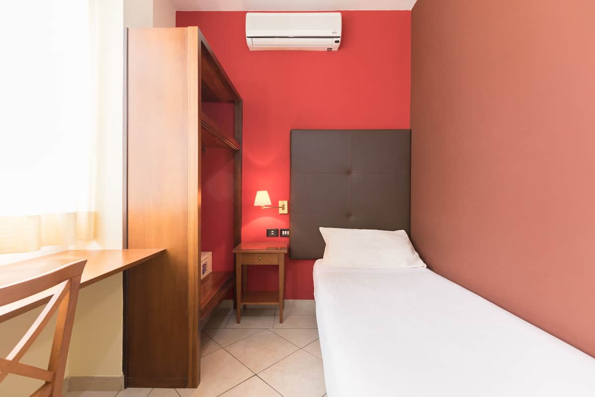 A hotel room with a bed, a desk, a chair, a nightstand with a lamp, and an air conditioner on the wall.