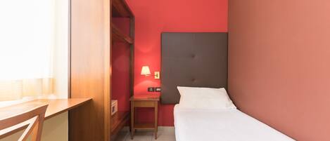 A hotel room with a bed, a desk, a chair, a nightstand with a lamp, and an air conditioner on the wall.