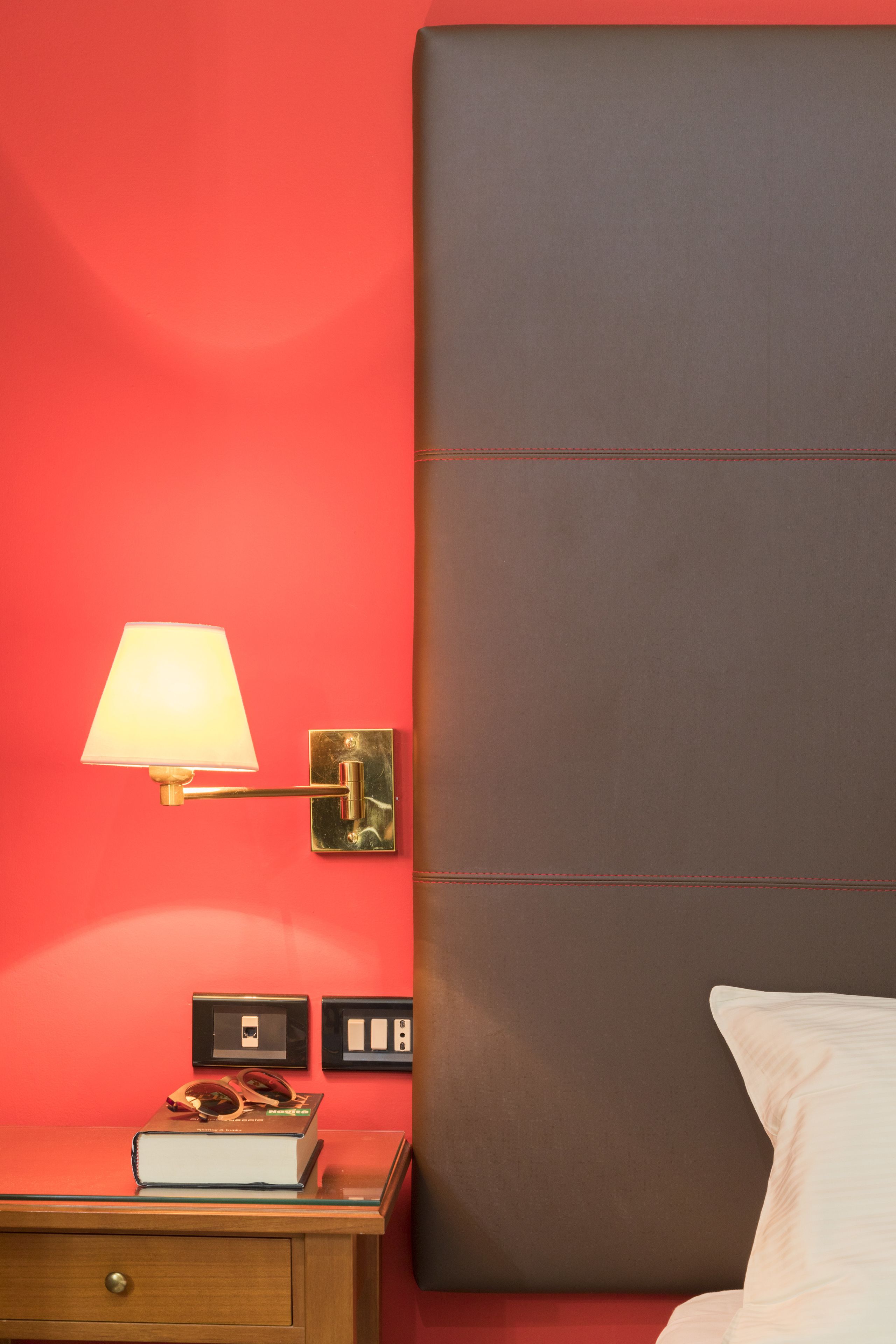 Une chambre d’hôtel avec un mur rouge, une lampe de chevet, une table de nuit avec un livre et des lunettes, et un lit avec un oreiller blanc.