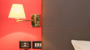Une chambre d’hôtel avec un mur rouge, une lampe de chevet, une table de nuit avec un livre et des lunettes, et un lit avec un oreiller blanc.