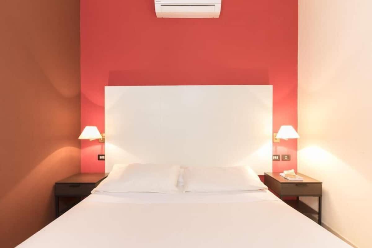 A hotel room with a large bed, two bedside tables with lamps, and an air conditioning unit on the wall.
