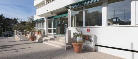 Un edificio moderno con fachada blanca, un toldo verde y una escalera que conduce a la entrada. Hay plantas en macetas cerca de la entrada y un extintor de incendios montado en la pared.