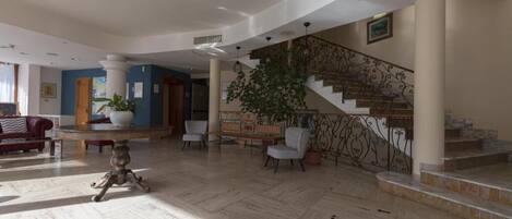 Un vestíbulo amplio con piso de mármol, una escalera con barandales de metal ornamentados y una zona de estar con una mesa y sillas.