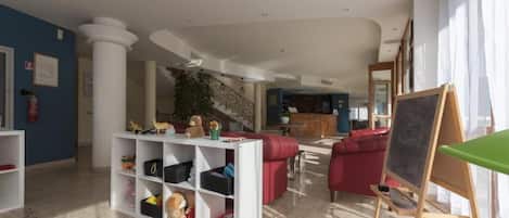 Un salón amplio con un sofá rojo, un estante blanco lleno de juguetes, una mesa verde y una pizarra.