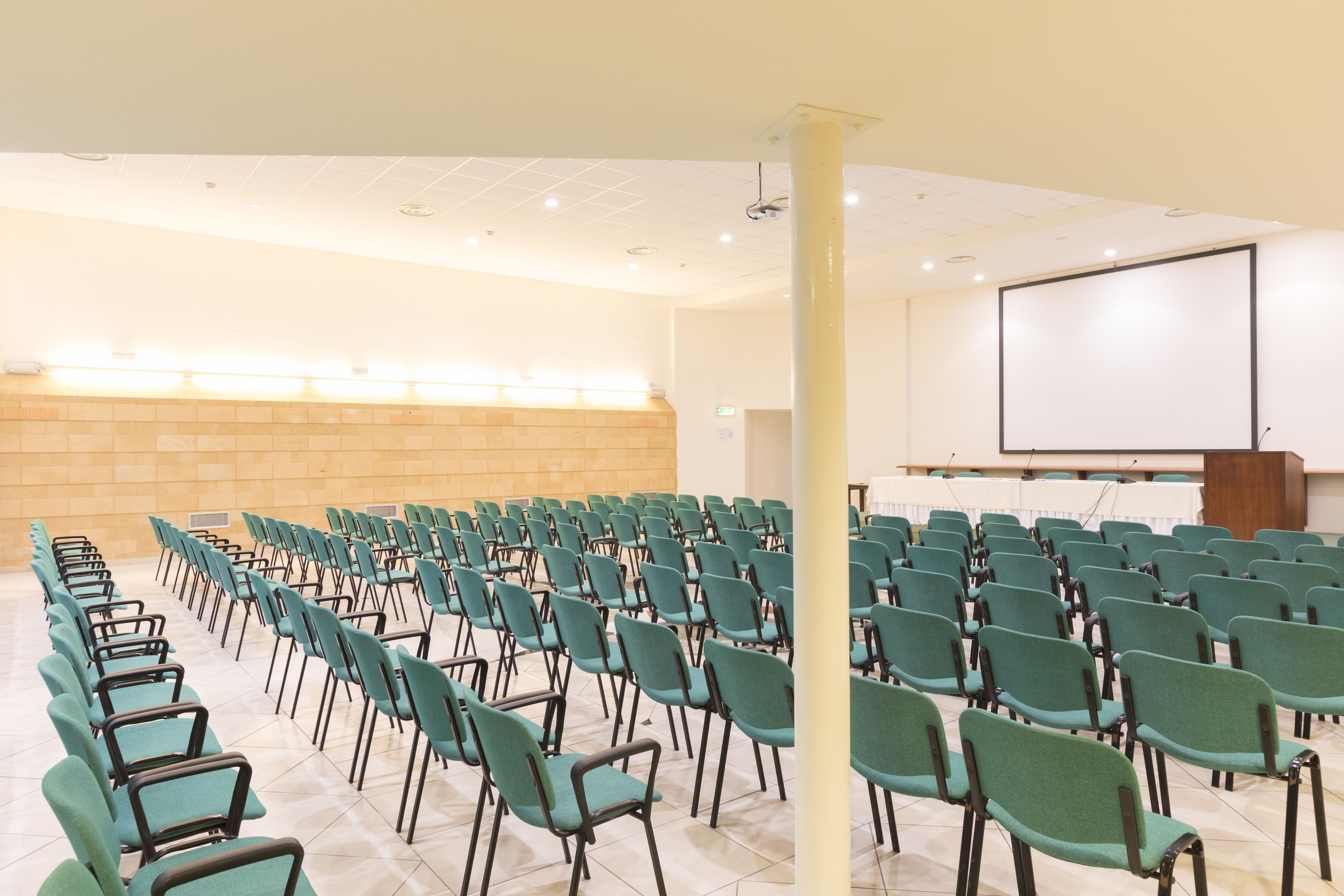 Un salón de conferencias con filas de sillas verdes y una pantalla de proyección blanca.