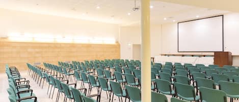 Un salón de conferencias con filas de sillas verdes y una pantalla de proyección blanca.