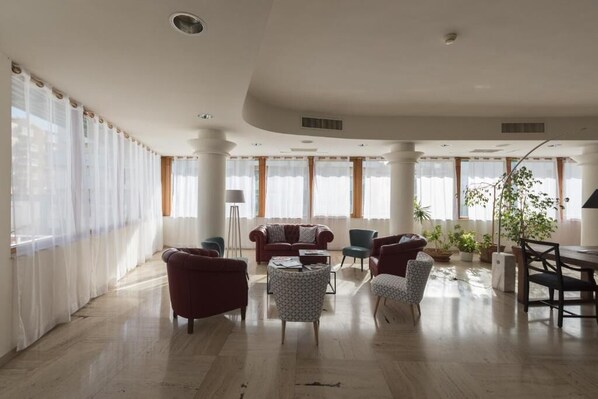 A spacious living room with a sofa set, a coffee table, and a dining table with chairs. There are large windows with curtains, a floor lamp, and several potted plants. - Hotel La Baia (Bari)