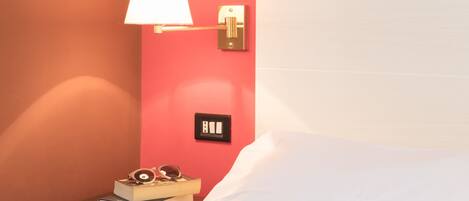 A neatly made bed with white linens, a bedside lamp, and a small table with a book and sunglasses.