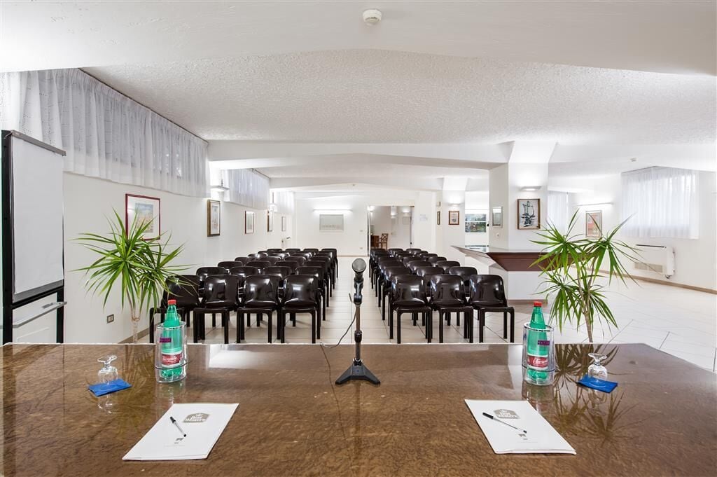 Un salón de conferencias con una larga mesa, sillas, un micrófono y dos plantas en macetas.