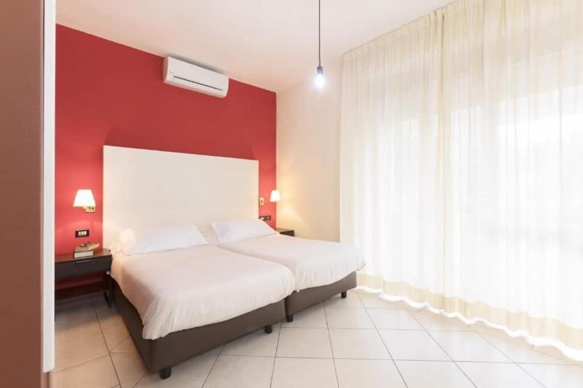 A double bed with a white headboard, two bedside tables with lamps, a red accent wall, and a large window with sheer curtains.