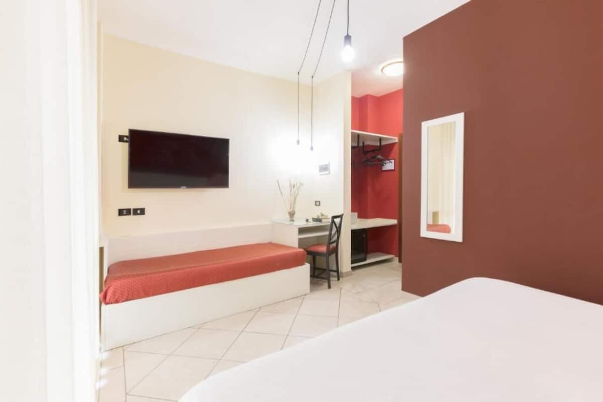 A compact hotel room with a bed, a desk, a chair, a TV mounted on the wall, and a mirror.