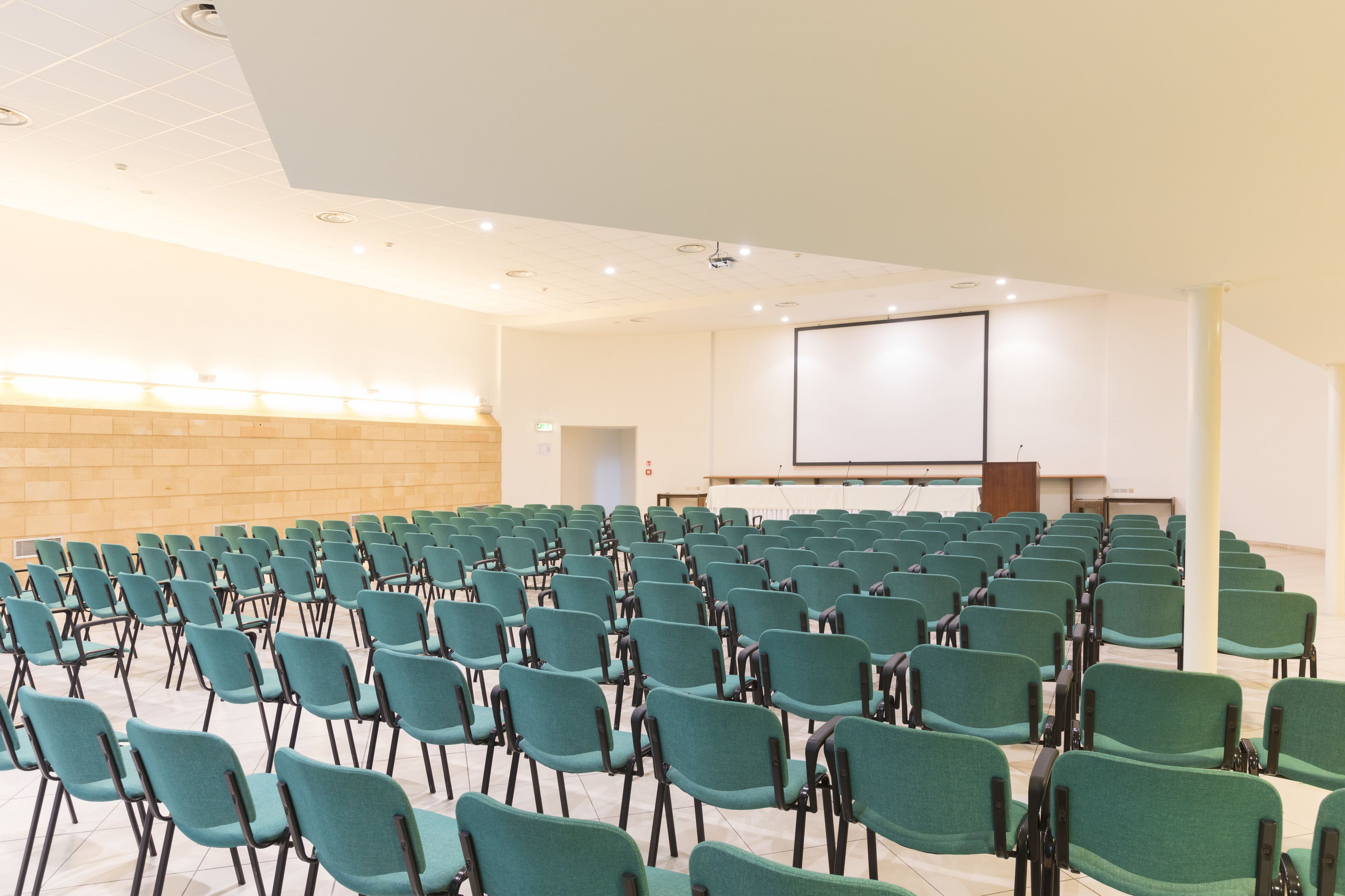 Une salle de conférence avec des rangées de chaises de couleur turquoise, un écran de projection et un pupitre.