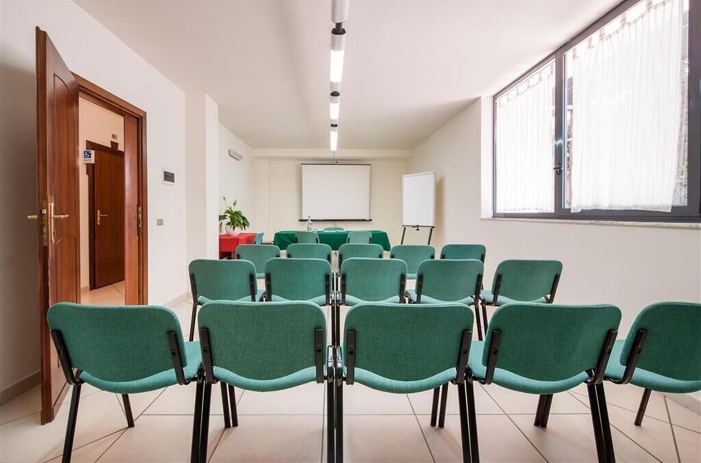 Un salón de conferencias con filas de sillas color turquesa, una pizarra y una pantalla para proyector.