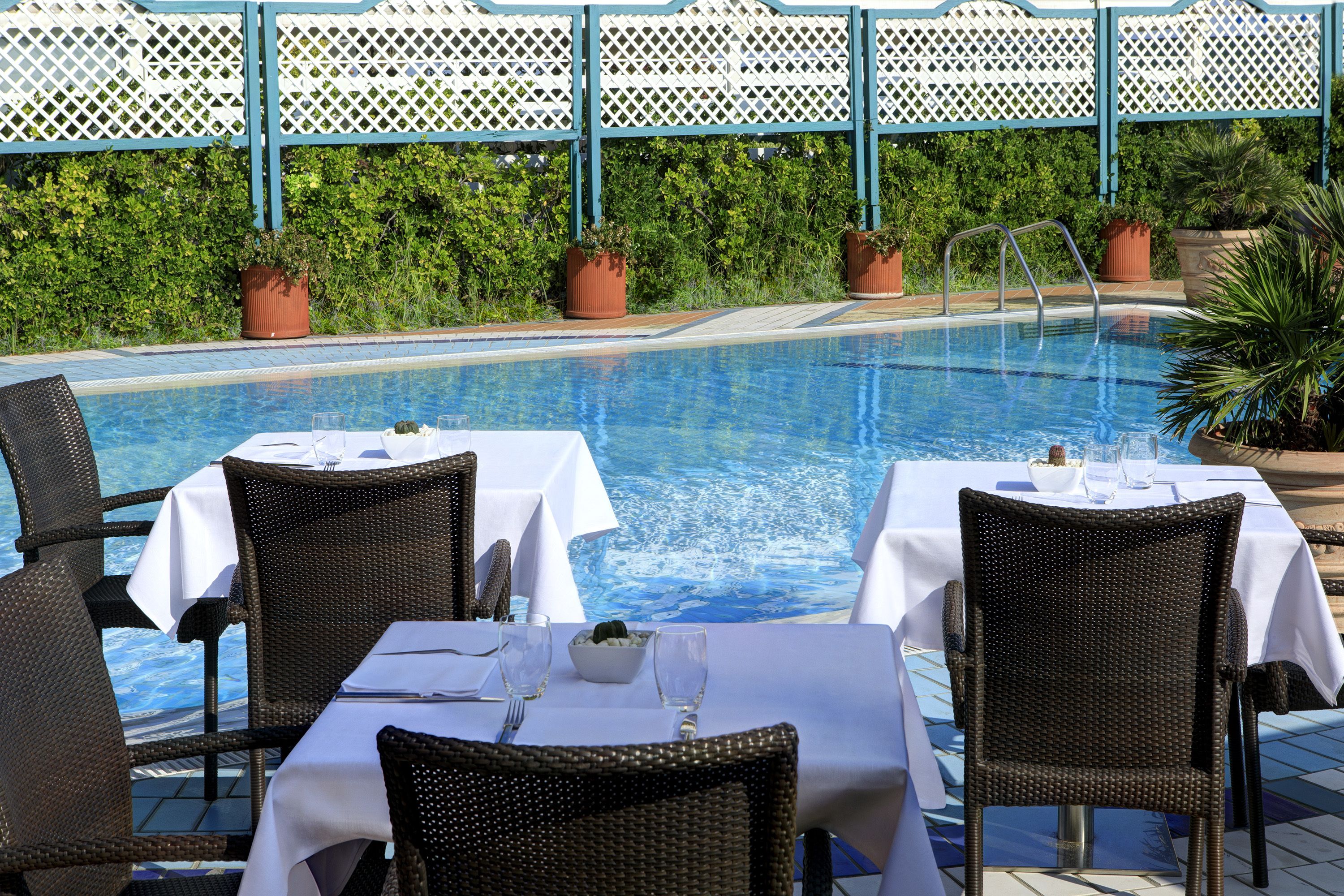 A poolside dining area with tables and chairs, a pool, and a white fence in the background.