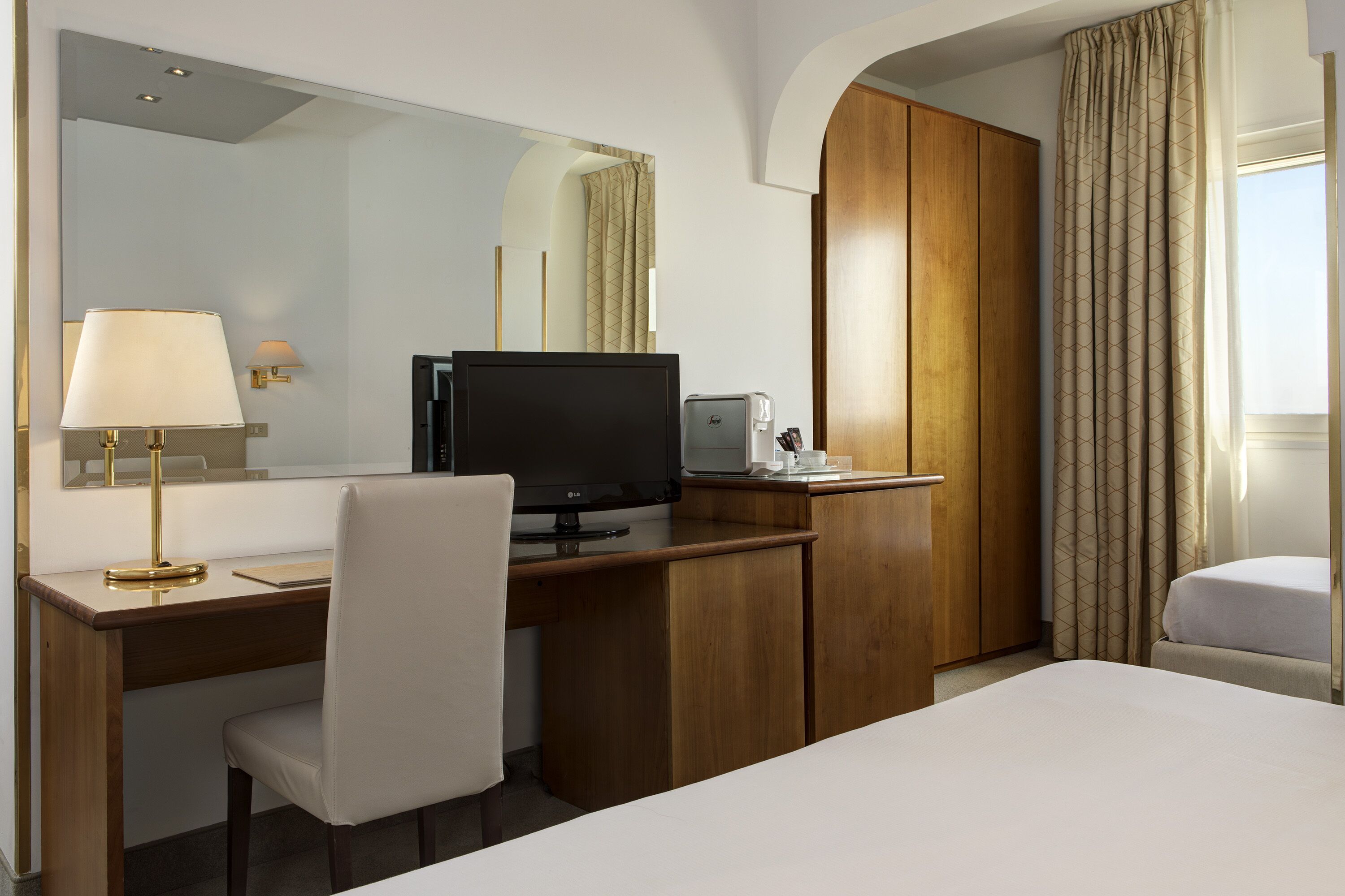 A hotel room with a wooden desk, a computer, a lamp, a bed, a wardrobe, and a window with curtains.