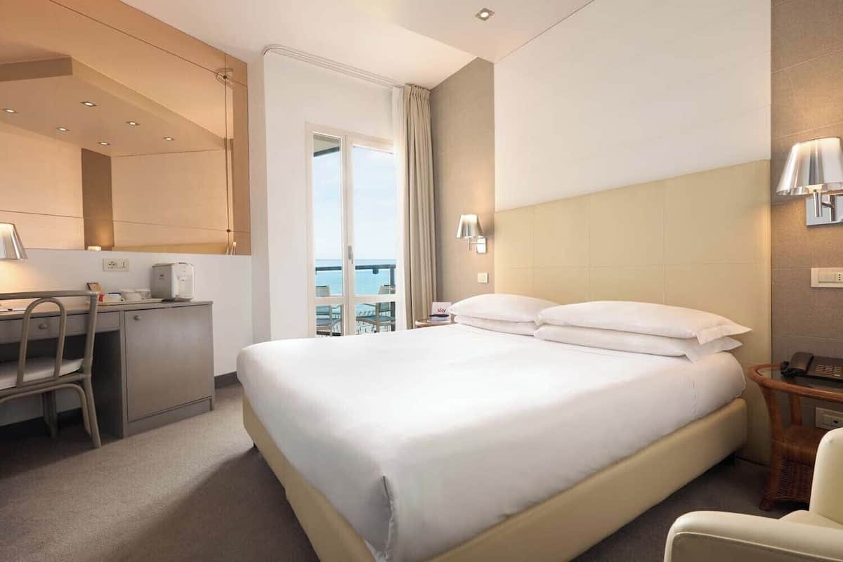 Een hotelkamer met een bed, een bureau, een stoel, een lamp en uitzicht op zee.
