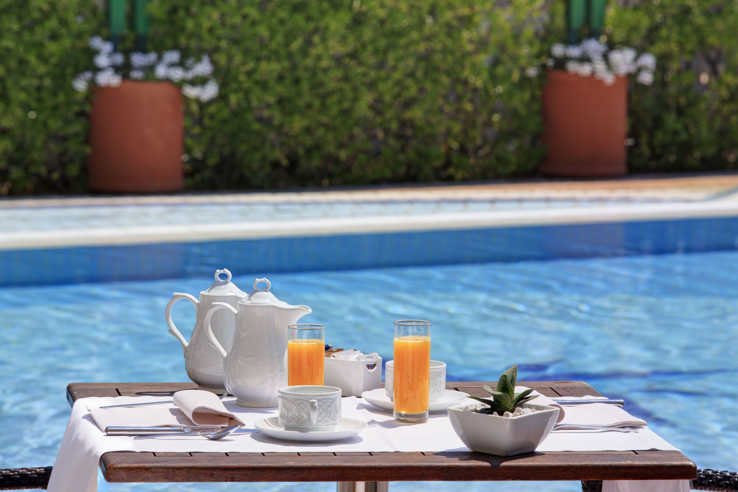 Une table dressée avec des aliments pour le petit-déjeuner, placée près d’une piscine, comprenant une théière, des tasses et un saladier rempli de fruits.