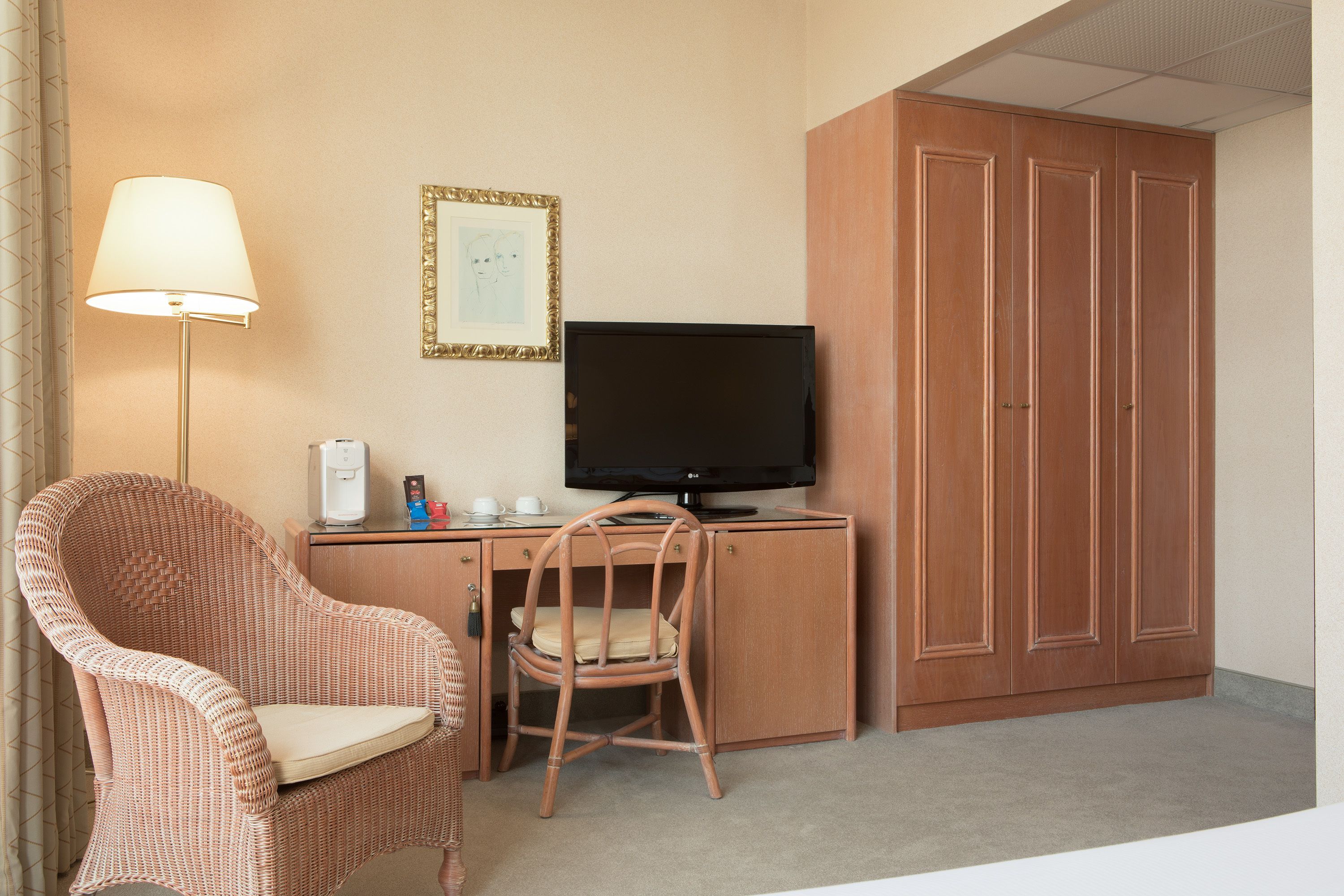 A hotel room with a wicker chair, a desk with a television, a coffee maker, and a wardrobe.