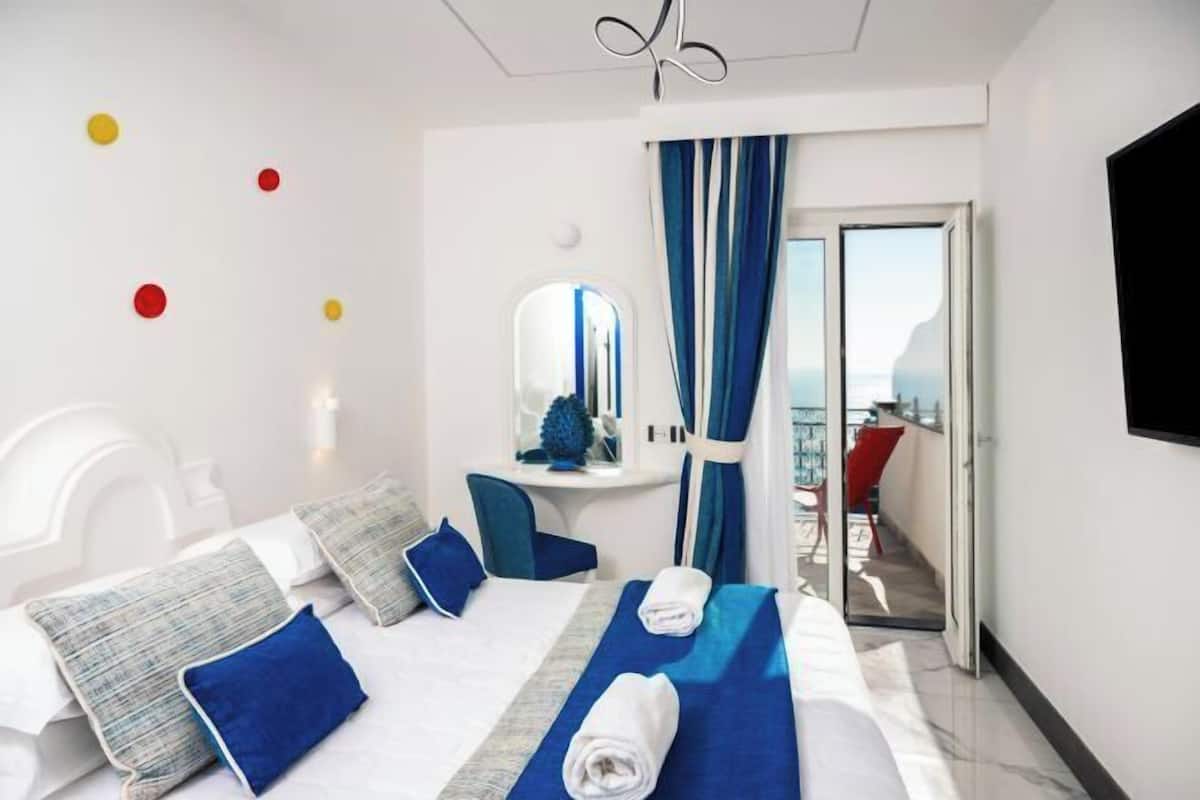 A hotel room with a bed, a desk, a chair, a mirror, and a balcony with a view.