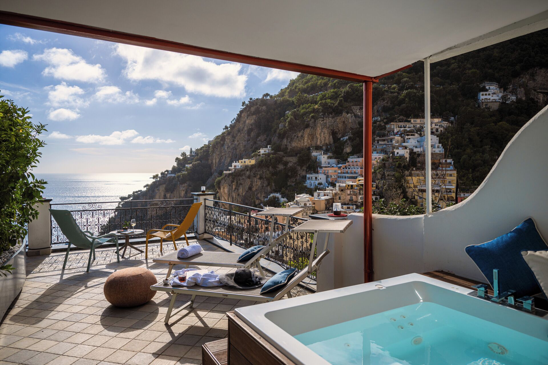 A balcony with a view of the sea, a jacuzzi, lounge chairs, and a dining table.