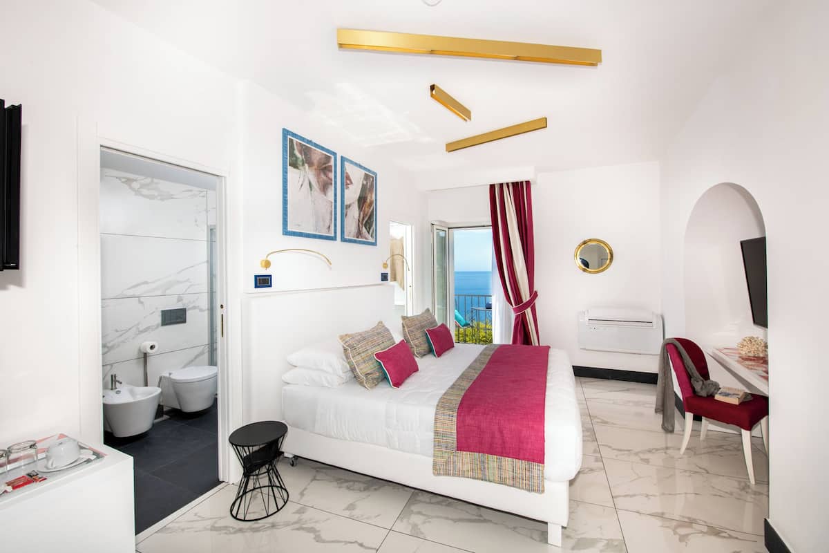 A modern hotel room with a bed, a desk, a chair, a bathroom, and a view of the sea.