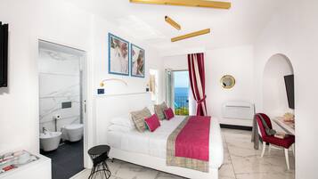 A modern hotel room with a bed, a desk, a chair, a bathroom, and a view of the sea.