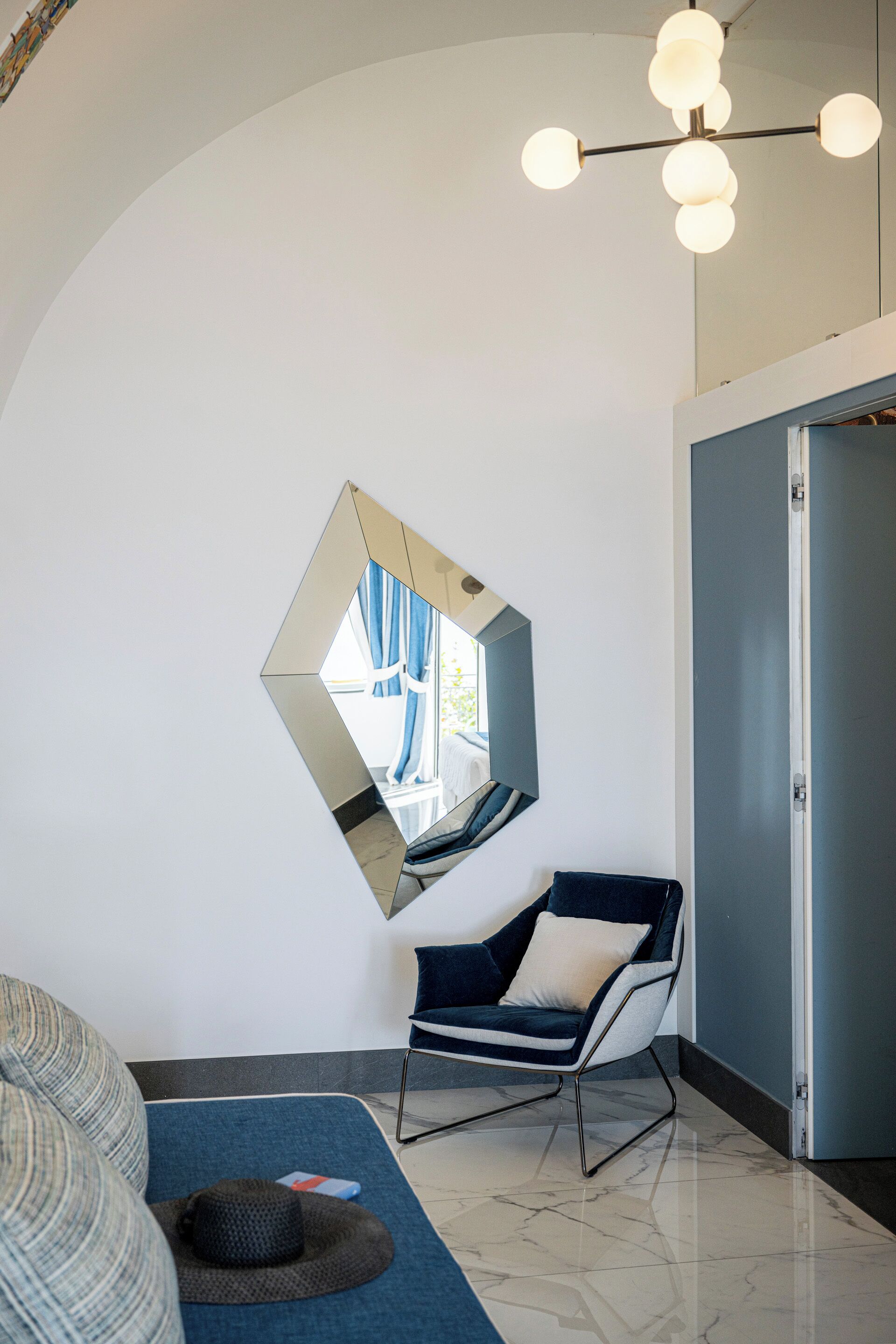 Una habitación moderna con un sofá azul, una silla, un espejo de pared original y una lámpara en el techo.