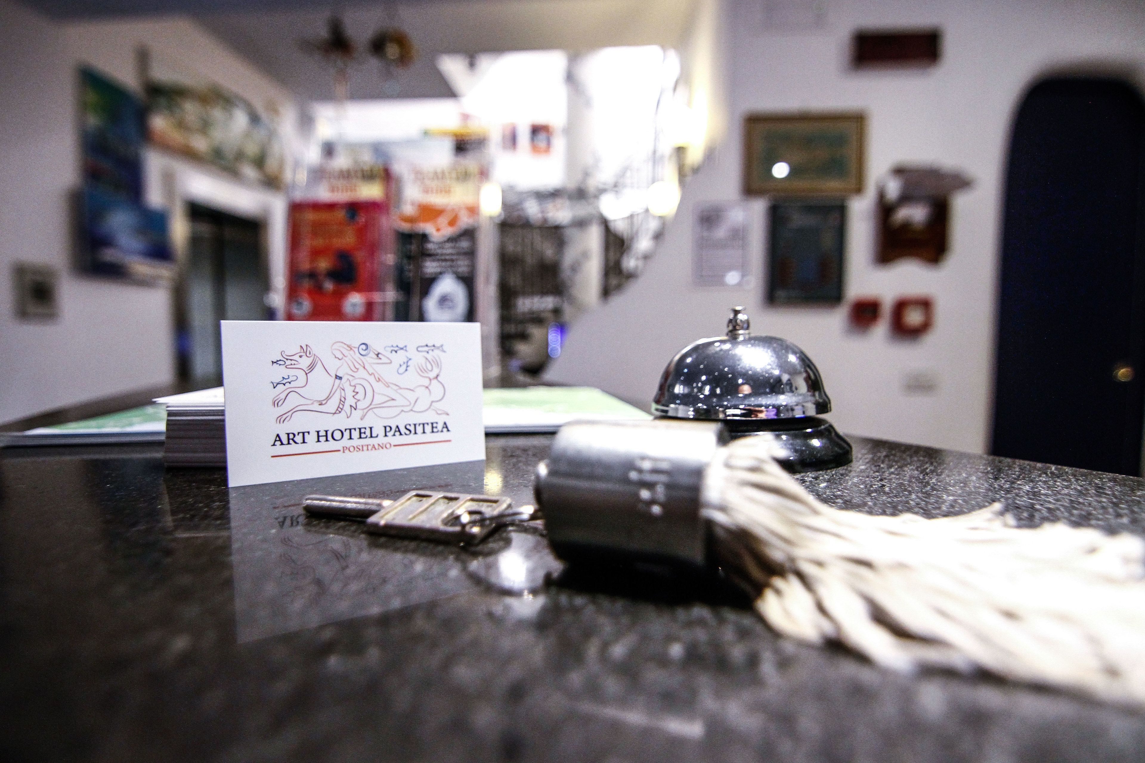 A hotel reception desk with a sign reading "ART HOTEL PASITEA POSTAN 2020," keys, a camera, and a bell.