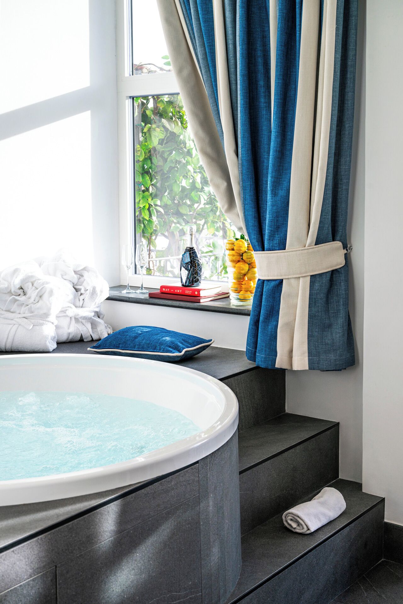 Um banheiro moderno com hidromassagem, degraus revestidos de azulejos escuros, uma janela com cortinas e vista para o verde lá fora.