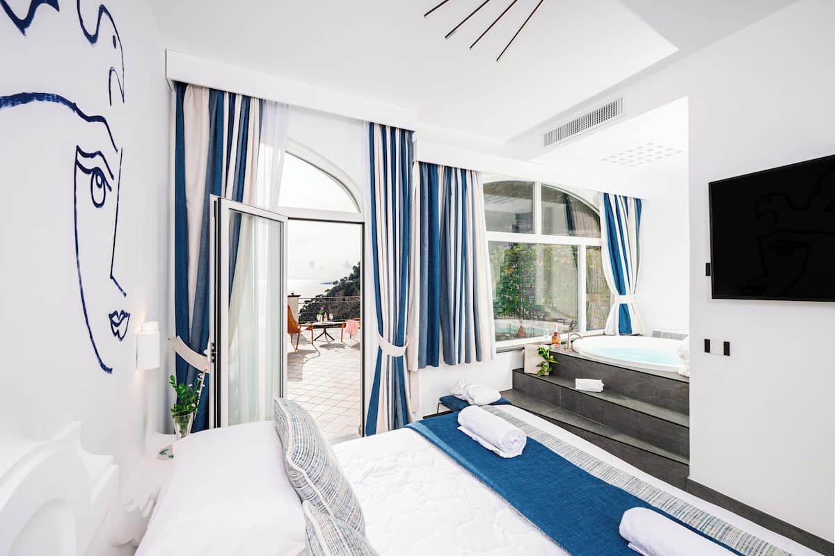 A modern hotel room with a large bed, a view of the outdoors, a seating area with a table and chairs, and a hot tub.