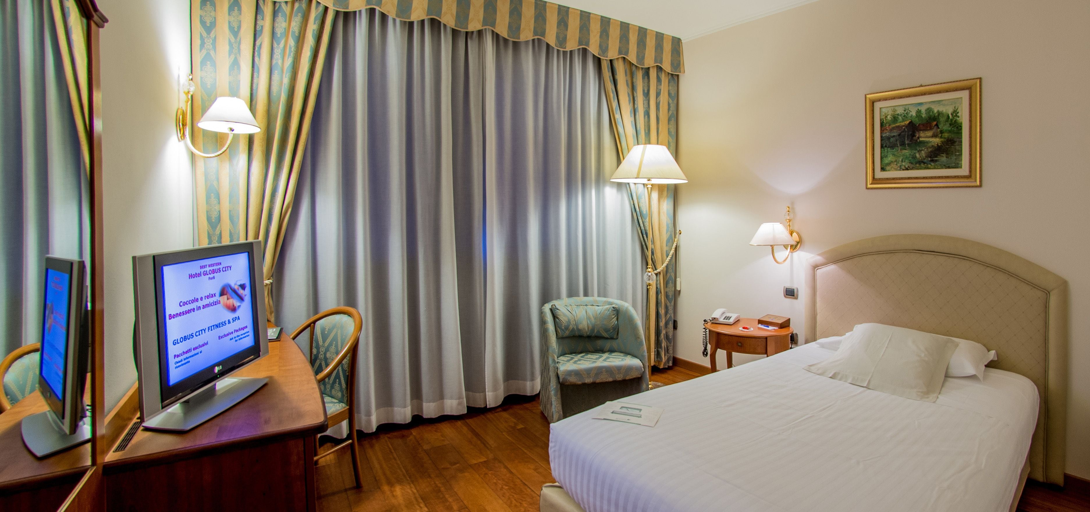 Une chambre d’hôtel avec un lit, un bureau avec un ordinateur, une chaise, une lampe et des rideaux.